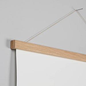 Puede incluir: Un colgador de póster de madera de color claro con una hoja de papel blanca adjunta. La barra de madera es rectangular y tiene una veta de madera natural. Una cuerda está unida a la parte superior de la barra de madera para colgarla.