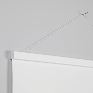 May include: A white wooden poster hanger with a white poster suspended from a thin wire against a gray wall. The top of the poster is secured by the hanger, which is attached to the wall with a wire.