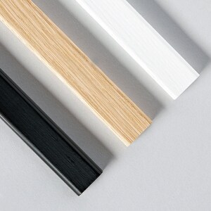 May include: Close-up of three wooden picture frame pieces in black, natural wood, and white. The frame pieces are angled diagonally across a light gray background, showcasing the different colors and textures of the wood.