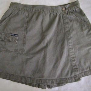 Può includere: Una gonna avvolgente color kaki con una grande tasca cargo sul lato sinistro. La gonna ha una chiusura a bottone e un'etichetta del marchio Johnny O.