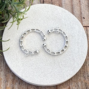 May include: Two silver-colored chain link charms, each in a semi-circular shape, are displayed on a light gray, circular surface. The charms are positioned on a wooden surface, with a green plant in the background.
