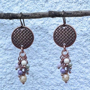 May include: A pair of copper-colored earrings. Each earring features a woven disc design, connected to a cluster of small, colorful beads in shades of cream, lavender, and green. The earrings are suspended from a simple hook.