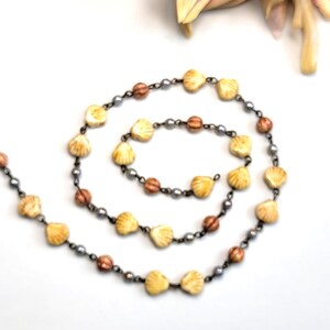 May include: A necklace featuring a repeating pattern of cream-colored shell-shaped beads, small metallic grey spherical beads, and larger round brown textured beads connected by a thin dark metal chain, arranged in a spiral shape on a white surface.
