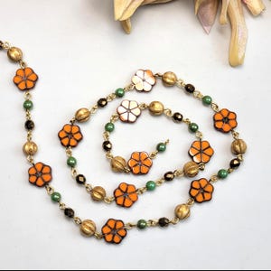 May include: A necklace featuring a gold-tone chain interspersed with orange flower-shaped beads, small green round beads, dark brown faceted beads, and textured gold-tone spherical beads arranged in a repeating pattern along the length of the chain.