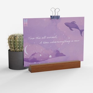 Puede incluir: Una impresión acuarela púrpura y rosa con una cita que dice "Dale la vuelta a todo, Un momento en el que todo es nuevo." La impresión presenta una luna creciente, estrellas y una ballena. La impresión se muestra en un caballete de madera.