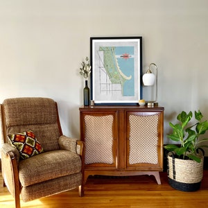 Może przedstawiać: Salon z meblami w stylu mid-century. Brązowy fotel z wzorzystą poduszką stoi obok drewnianej szafki z nadrukiem mapy. Złota lampa i roślina doniczkowa w plecionym koszu r&oacute;wnież są obecne. Pok&oacute;j ma drewniane podłogi i neutralne ściany.