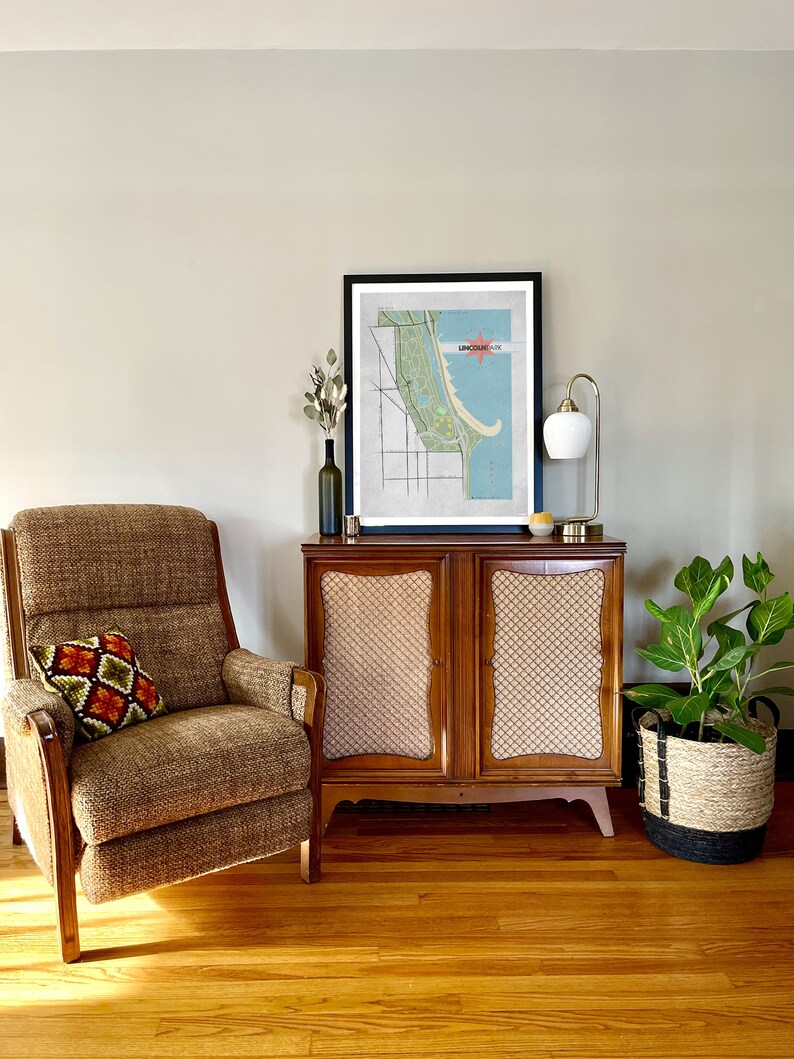 Może przedstawiać: Salon z meblami w stylu mid-century. Brązowy fotel z wzorzystą poduszką stoi obok drewnianej szafki z nadrukiem mapy. Złota lampa i roślina doniczkowa w plecionym koszu r&oacute;wnież są obecne. Pok&oacute;j ma drewniane podłogi i neutralne ściany.