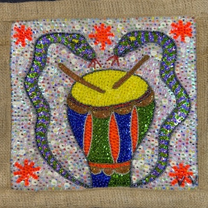 May include: A square, beaded artwork featuring a colorful drum with drumsticks, flanked by two green and purple snakes. The background is covered in sequins, with orange star-shaped accents. The artwork is set against a burlap background.