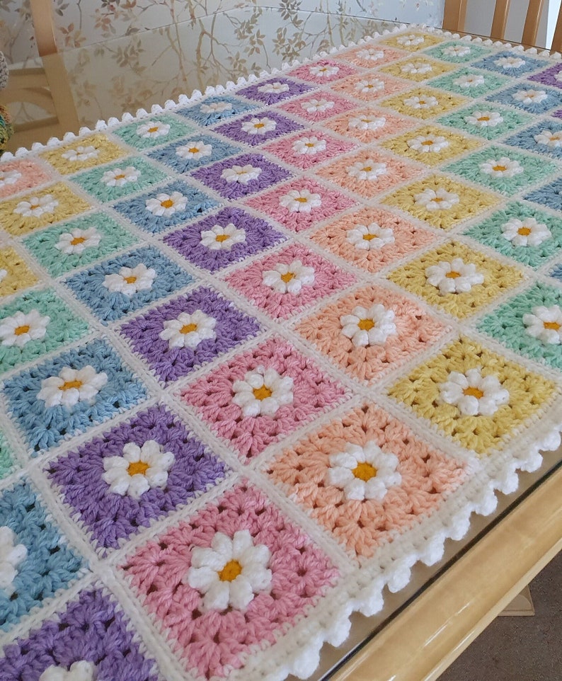 May include: A crocheted baby blanket with a rainbow of colors and white daisy flowers. The blanket has a white crocheted border.