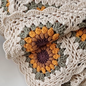 May include: A crocheted blanket with a sunflower design in shades of brown, orange, and green. The blanket is made with a cream-colored yarn and has a scalloped edge.