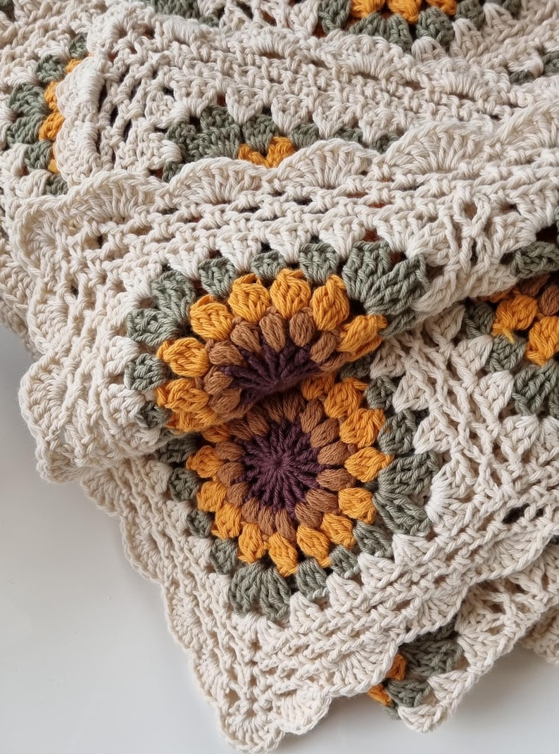 May include: A crocheted blanket with a sunflower design in shades of brown, orange, and green. The blanket is made with a cream-colored yarn and has a scalloped edge.