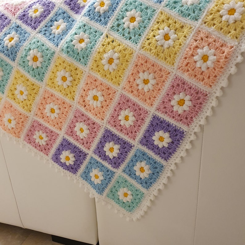 May include: A crocheted baby blanket with a daisy pattern in pastel colors, including pink, yellow, blue, and purple. The blanket has a white crocheted border.