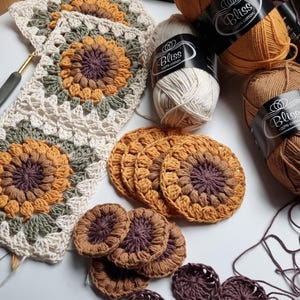 May include: A close-up of crocheted sunflower motifs in various shades of brown, yellow, and green. The motifs are arranged in a square pattern and are surrounded by balls of yarn labeled "Bliss 100% cotton".