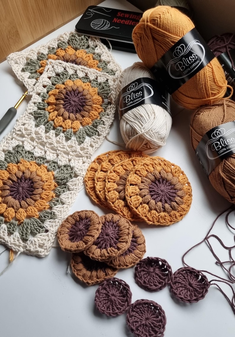 May include: A close-up of crocheted sunflower motifs in various shades of brown, yellow, and green. The motifs are arranged in a square pattern and are surrounded by balls of yarn labeled "Bliss 100% cotton".