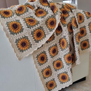 May include: A crocheted throw blanket with a sunflower pattern in shades of brown, yellow, and green. The blanket is made with a white yarn and has a scalloped edge.
