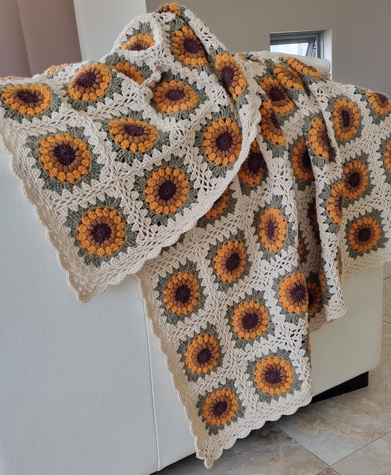 May include: A crocheted throw blanket with a sunflower pattern in shades of brown, yellow, and green. The blanket is made with a white yarn and has a scalloped edge.