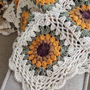 May include: A crocheted throw blanket with a sunflower pattern in shades of brown, yellow, and green. The blanket is made with a white yarn and has a scalloped edge.
