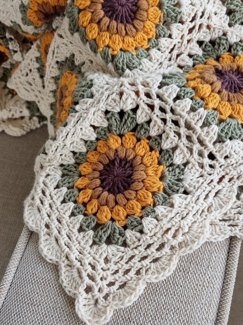 May include: A crocheted throw blanket with a sunflower pattern in shades of brown, yellow, and green. The blanket is made with a white yarn and has a scalloped edge.