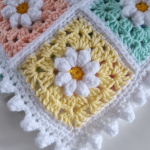 May include: A crocheted baby blanket with a white border and a daisy pattern in shades of yellow, pink, and green.