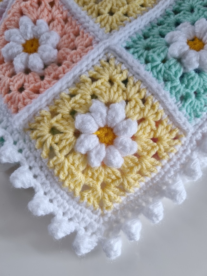 May include: A crocheted baby blanket with a white border and a daisy pattern in shades of yellow, pink, and green.