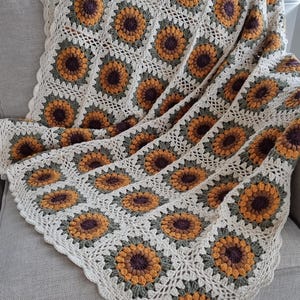 May include: A crocheted throw blanket with a sunflower pattern in shades of brown, yellow, and green. The blanket is made with a white yarn and has a scalloped edge.