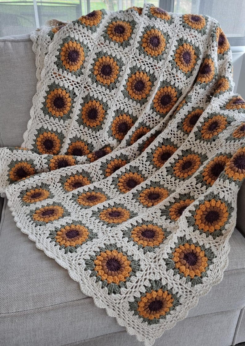 May include: A crocheted throw blanket with a sunflower pattern in shades of brown, yellow, and green. The blanket is made with a white yarn and has a scalloped edge.