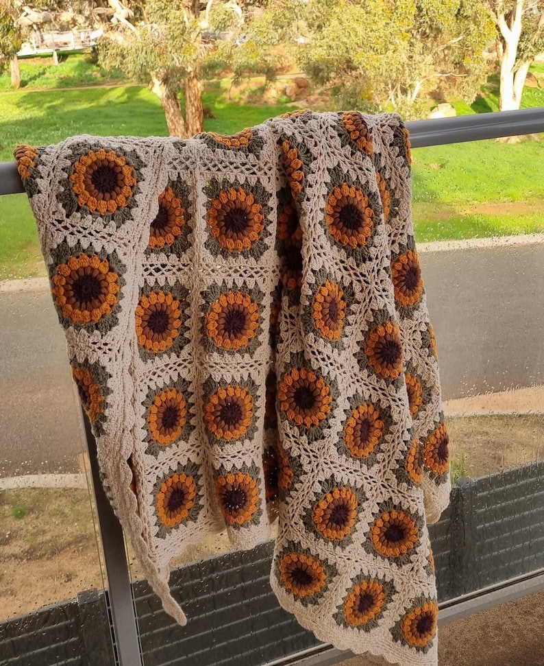 May include: A crocheted throw blanket with a sunflower pattern in shades of brown, yellow, and green. The blanket is draped over a railing.