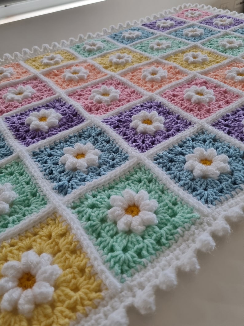 May include: A crocheted baby blanket with a white border and a daisy pattern in pastel colors, including yellow, green, blue, purple, pink, and orange.