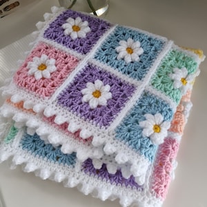 May include: A crocheted baby blanket with a daisy pattern in pastel colors, including pink, purple, blue, and green. The blanket has a white crocheted border.