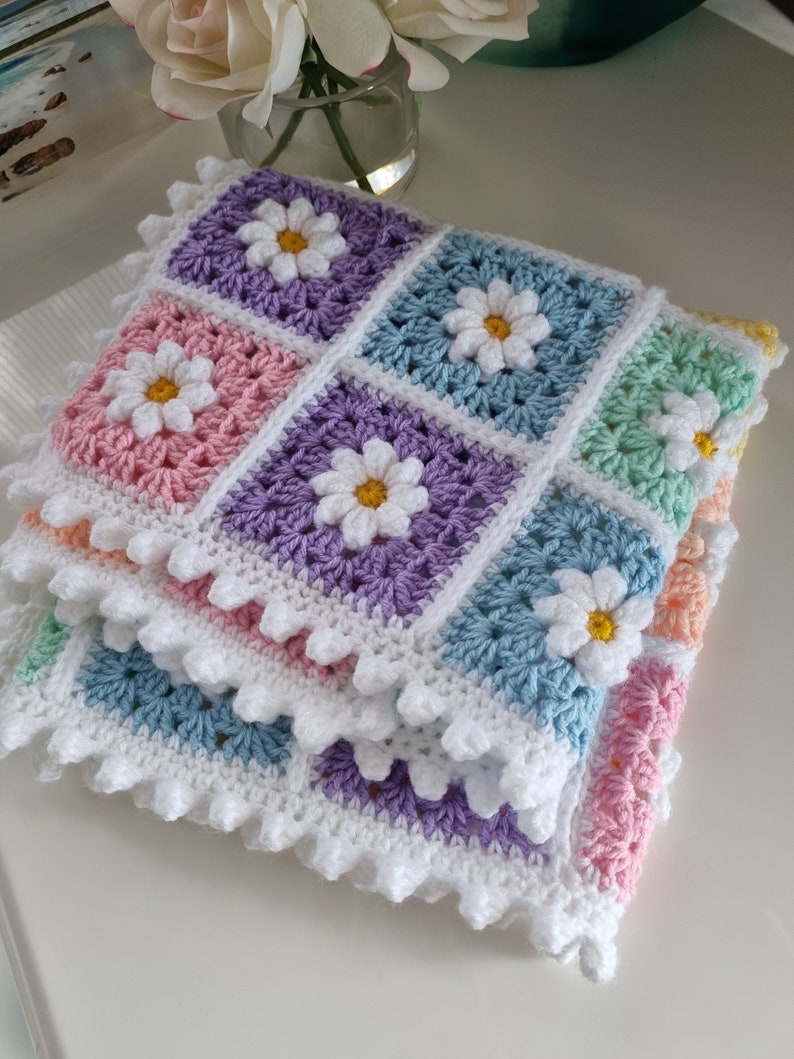 May include: A crocheted baby blanket with a daisy pattern in pastel colors, including pink, purple, blue, and green. The blanket has a white crocheted border.