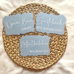 Könnte beinhalten: Drei hellblaue Acrylschilder mit weißer Schrift auf einem gewebten Platzset. Die Schilder lauten "Open Bar Cheers", "Gästebuch Bitte Verewigt Euch" und "Notfallkörbchen Bitte Bedient Euch".