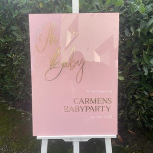 Könnte beinhalten: Ein rosa Schild mit goldfarbenem Schriftzug "Oh Baby" und "Carmens Babyparty" mit dem Datum "18. Mai 2025". Das Schild wird auf einer weißen Staffelei vor grünem Laub ausgestellt.