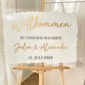 Könnte beinhalten: Ein weißes Acrylschild mit goldener Beschriftung, auf dem "Willkommen zu unserer Hochzeit Julia & Alexander 13. Juli 2024" steht. Das Schild steht auf einem Holzständer.