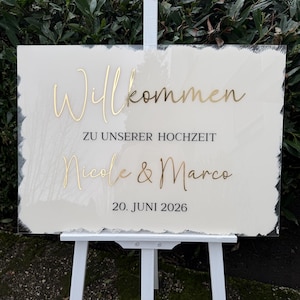 Könnte beinhalten: Ein weißes Hochzeits-Willkommensschild aus Acryl mit goldfarbenen Buchstaben. Auf dem Schild steht "Willkommen zu unserer Hochzeit Nicole & Marco 20. JUNI 2026" und es wird auf einer weißen Staffelei präsentiert.