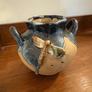 May include: A handcrafted ceramic vase with a round body and two handles. The vase features a dragonfly applique and is glazed in shades of blue, brown, and tan. The vase is a decorative item.