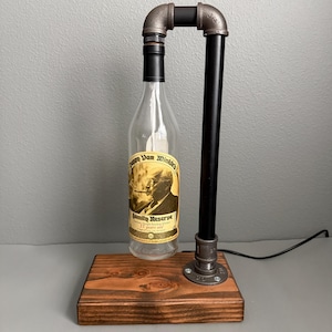 K&ouml;nnte beinhalten: Eine umfunktionierte Flaschenlampe mit einer durchsichtigen Glasflasche und einem Vintage-Etikett. Die Lampe hat einen schwarzen Rohrahmen und steht auf einem gebeizten Holzsockel. Die Lampe enth&auml;lt ein Netzkabel.