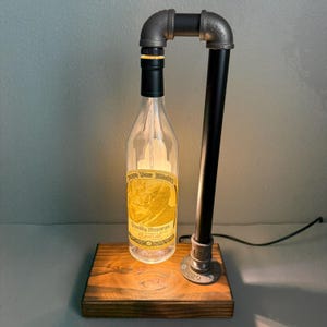 K&ouml;nnte beinhalten: Eine einzigartige Lampe im Industriestil aus einer wiederverwendeten Spirituosenflasche. Die klare Glasflasche hat ein Etikett mit Text und wird von innen beleuchtet. Die Lampe wird von schwarzen und grauen Metallrohren getragen und steht auf einem Holzsockel.
