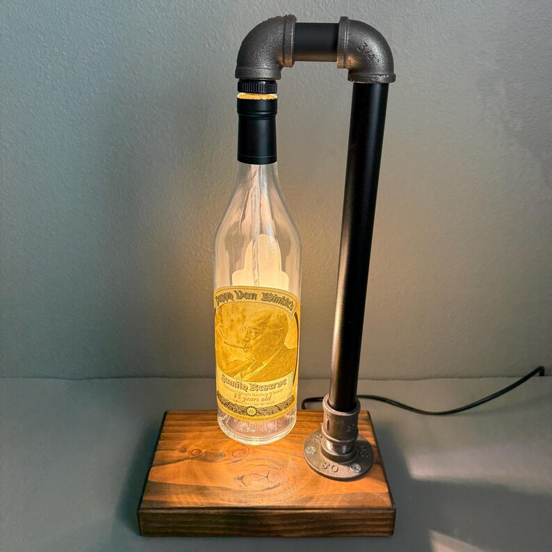 K&ouml;nnte beinhalten: Eine einzigartige Lampe im Industriestil aus einer wiederverwendeten Spirituosenflasche. Die klare Glasflasche hat ein Etikett mit Text und wird von innen beleuchtet. Die Lampe wird von schwarzen und grauen Metallrohren getragen und steht auf einem Holzsockel.