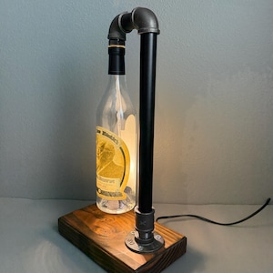 K&ouml;nnte beinhalten: Eine einzigartige Tischlampe aus einer recycelten, klaren Glasflasche, mit einem schwarzen Metallrohrrahmen und einem Holzsockel. Die Flasche hat ein Etikett mit goldenen und braunen Akzenten. Die Lampe ist beleuchtet und strahlt ein warmes Licht aus.