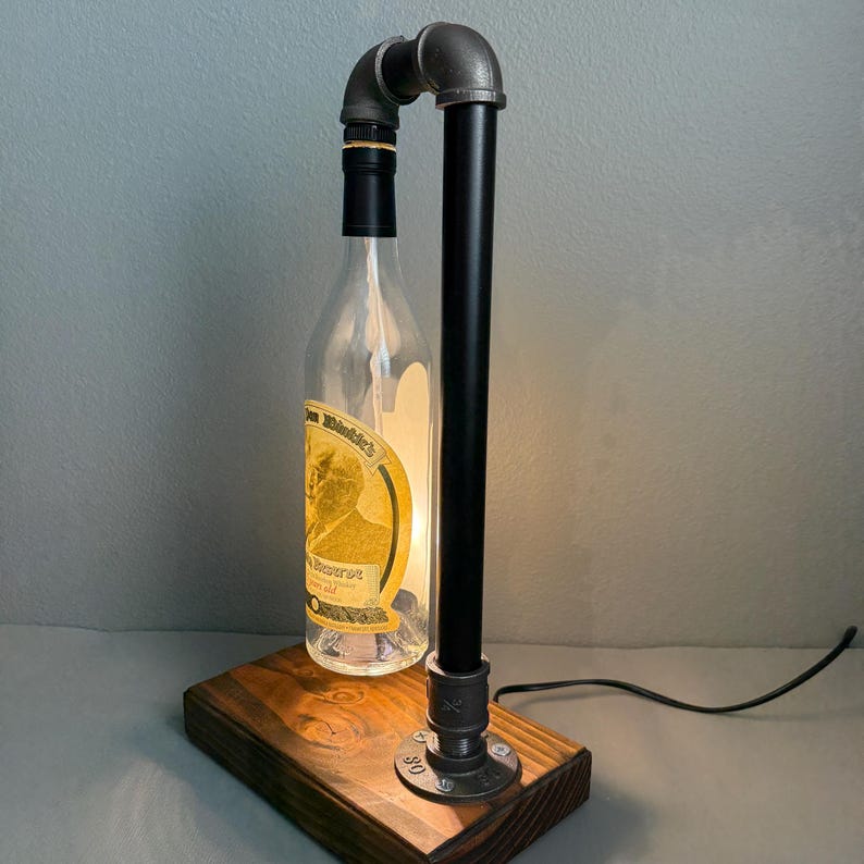K&ouml;nnte beinhalten: Eine einzigartige Tischlampe aus einer recycelten, klaren Glasflasche, mit einem schwarzen Metallrohrrahmen und einem Holzsockel. Die Flasche hat ein Etikett mit goldenen und braunen Akzenten. Die Lampe ist beleuchtet und strahlt ein warmes Licht aus.