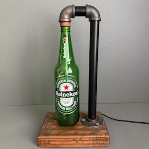 May include: A table lamp constructed from a green Heineken glass bottle, a black metal pipe frame, and a rectangular wooden base. The bottle features the Heineken Original Premium Malt Lager label with a red star. A black power cord extends from the base.