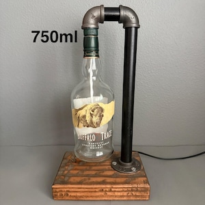May include: A lamp constructed from a 750ml Buffalo Trace Kentucky Straight Bourbon Whiskey bottle, featuring a dark metal pipe frame attached to a rectangular wooden base. The bottle has a clear glass body with a label depicting a buffalo.