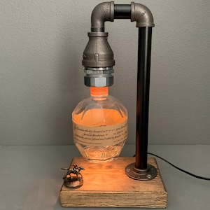 May include: A rustic table lamp made from black pipe fittings and a clear glass bottle. The bottle is illuminated from within, casting a warm glow. A small horse figurine sits on the wooden base.