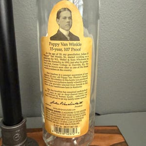 K&ouml;nnte beinhalten: Eine klare Glasflasche Pappy Van Winkle Bourbon mit einem gelben Etikett. Das Etikett zeigt ein Portr&auml;t eines Mannes und Text, der das Alter, den Alkoholgehalt und die Geschichte des Bourbons beschreibt. Die Flasche ist teilweise sichtbar.