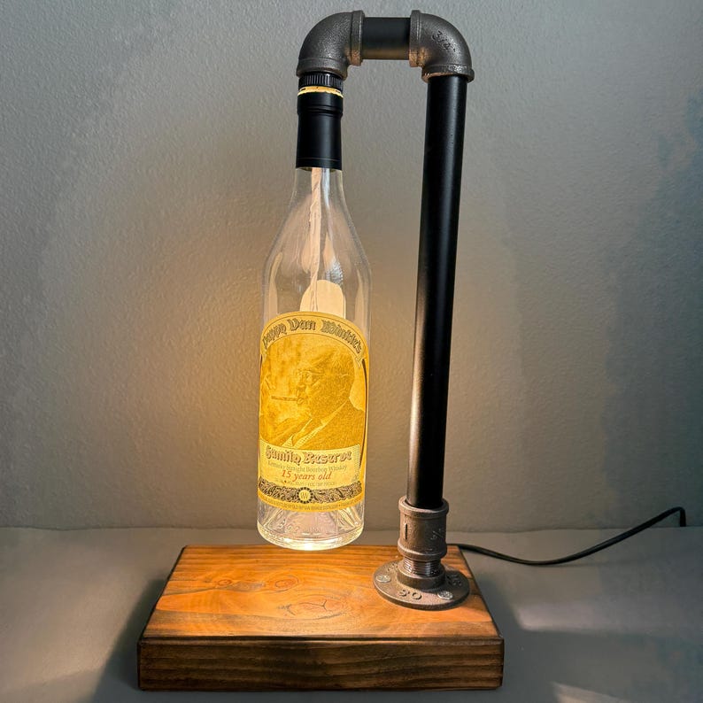 K&ouml;nnte beinhalten: Eine einzigartige Lampe aus einer wiederverwendeten Flasche, schwarzen Rohren und einem Holzsockel. Die Flasche hat ein Etikett mit der Aufschrift "Don Whitaker Family Reserve 15 years old". Die Lampe ist eingesteckt und leuchtet.