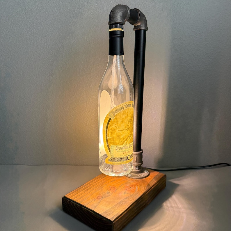 K&ouml;nnte beinhalten: Eine einzigartige Tischlampe aus einer recycelten Glasflasche, schwarzem Rohr und einem Holzsockel. Die Flasche hat einen schwarzen Verschluss und ein goldenes Etikett. Die Lampe ist beleuchtet und strahlt ein warmes Licht aus. Der Sockel besteht aus gebeiztem Holz.