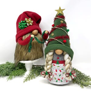 May include: Two Christmas gnomes. One gnome wears a red hat with green and gold bows, a brown beard, and red arms holding green garland. The other gnome is dressed as a Christmas tree with a gold star, red garland, and a white apron with Christmas lights.