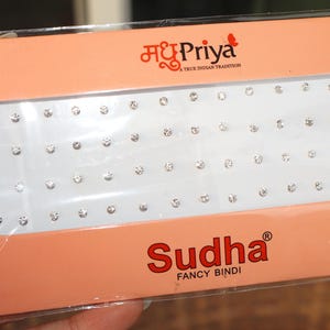 May include: A package of Sudha Fancy Bindi, featuring a rectangular tray filled with small, sparkling rhinestones. The packaging is peach-colored with the Madhu Priya logo and text in red.