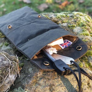 Könnte beinhalten: Eine dunkelgrüne gewachste Canvas-Tasche, geöffnet, um einen kleinen Union Jack-Aufnäher und ein Metallwerkzeug zu zeigen. Die Tasche ist mit Messingdruckknöpfen verschlossen und hat ein Lederinnenfutter. Die Tasche liegt auf einem moosbewachsenen Felsen.