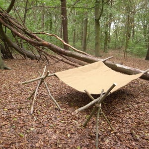May include: A tan canvas hammock supported by a wooden frame, situated in a forest setting. The frame is made from tied branches. The ground is covered in fallen leaves, with trees in the background.